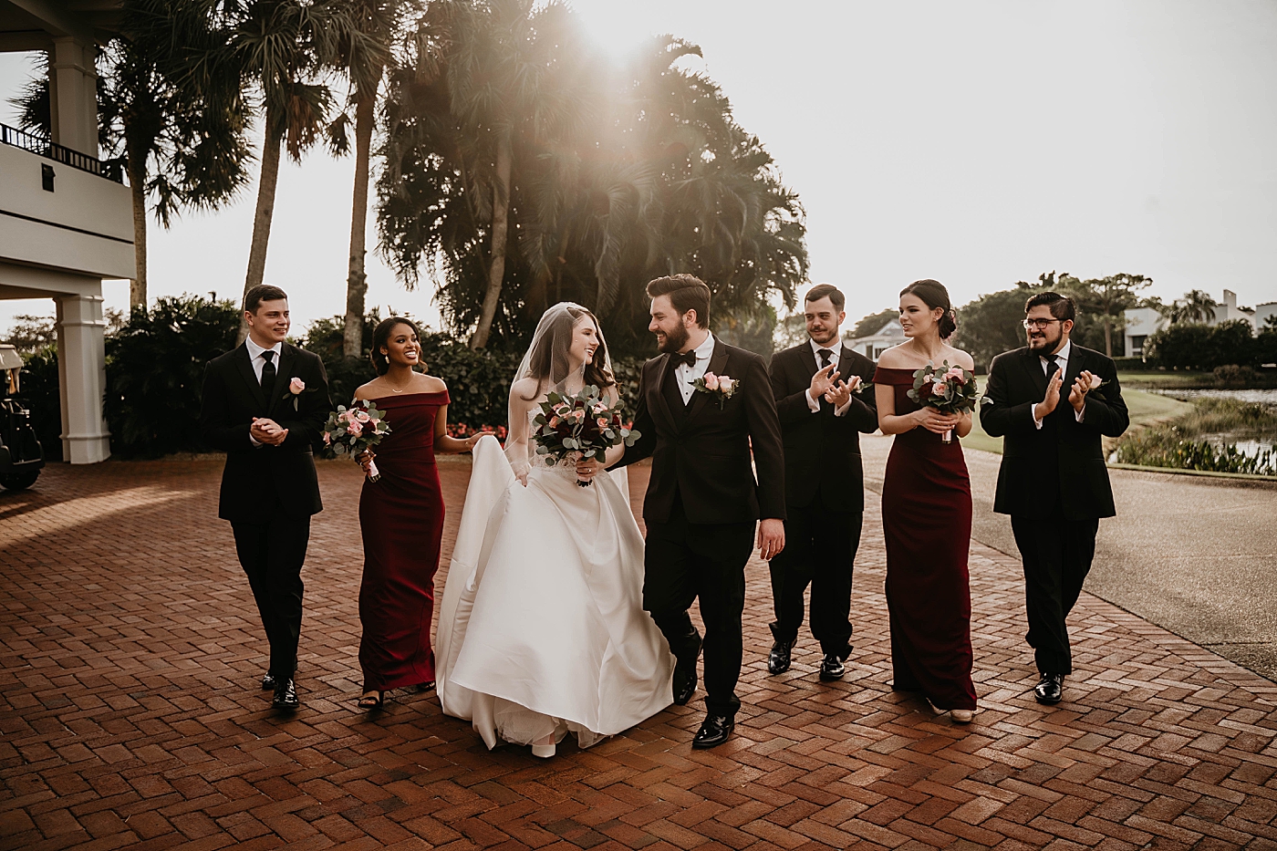 Bride and Groom walking holding hands and red pin white bouquet with wedding party behind them celebrating with sunset lens flare and palm trees Romantic Winter Wedding captured NYC Wedding Planner Poppy and Lynn