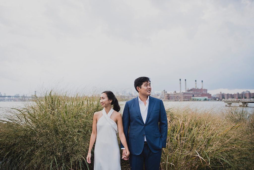 Couple holding hands and looking in opposite directions in front of grassy marsh by Hudson River