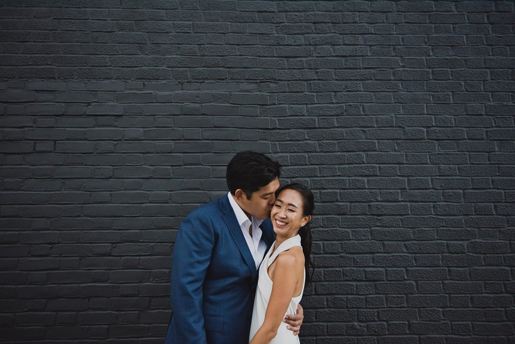 Groom kissing Bride on the cheek in front of black paint brick wall