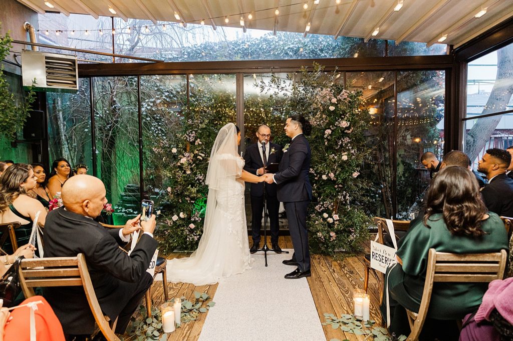 Bride and Groom holding hands for Ceremony with greenery around them and Officiant and guests watching