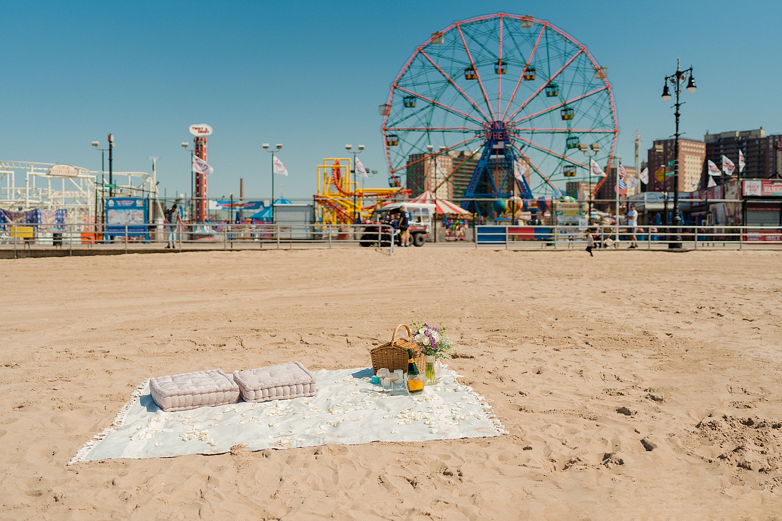 beach picnic proposal set up wide shot with pillows picnic basket blanket champagne and ferris wheel in background