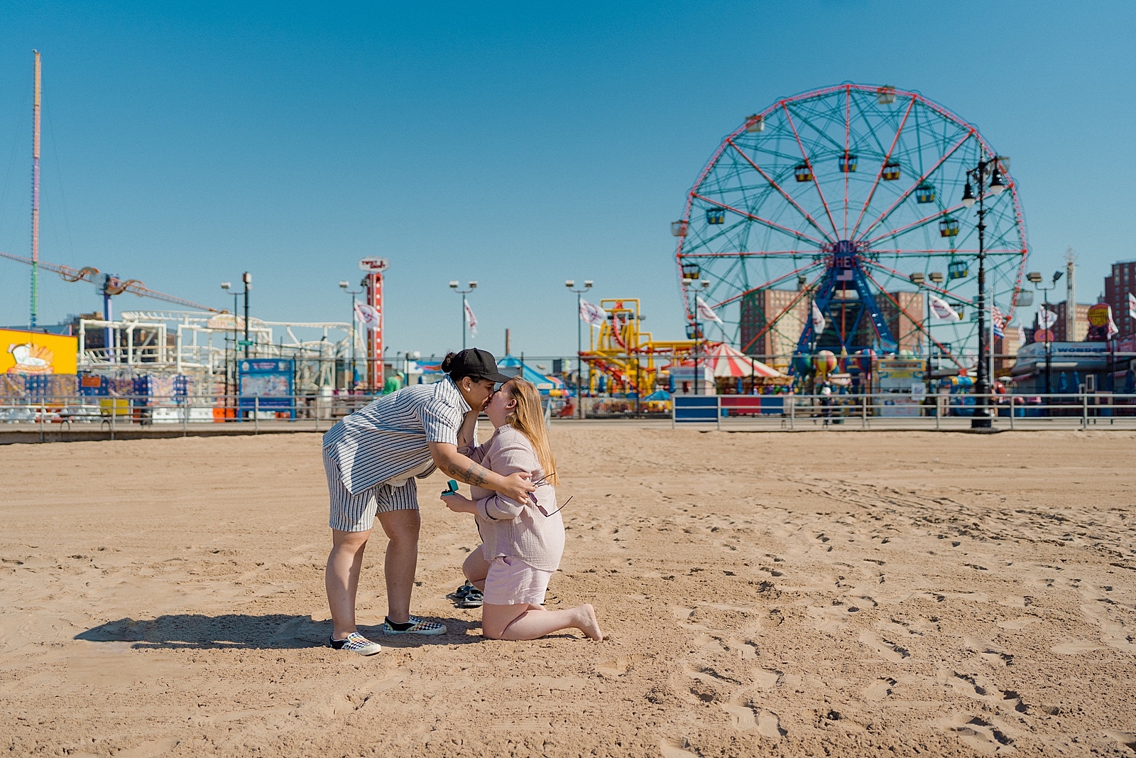 one woman proposing to other woman in front of coney island wonder wheel kissing with ring in hand