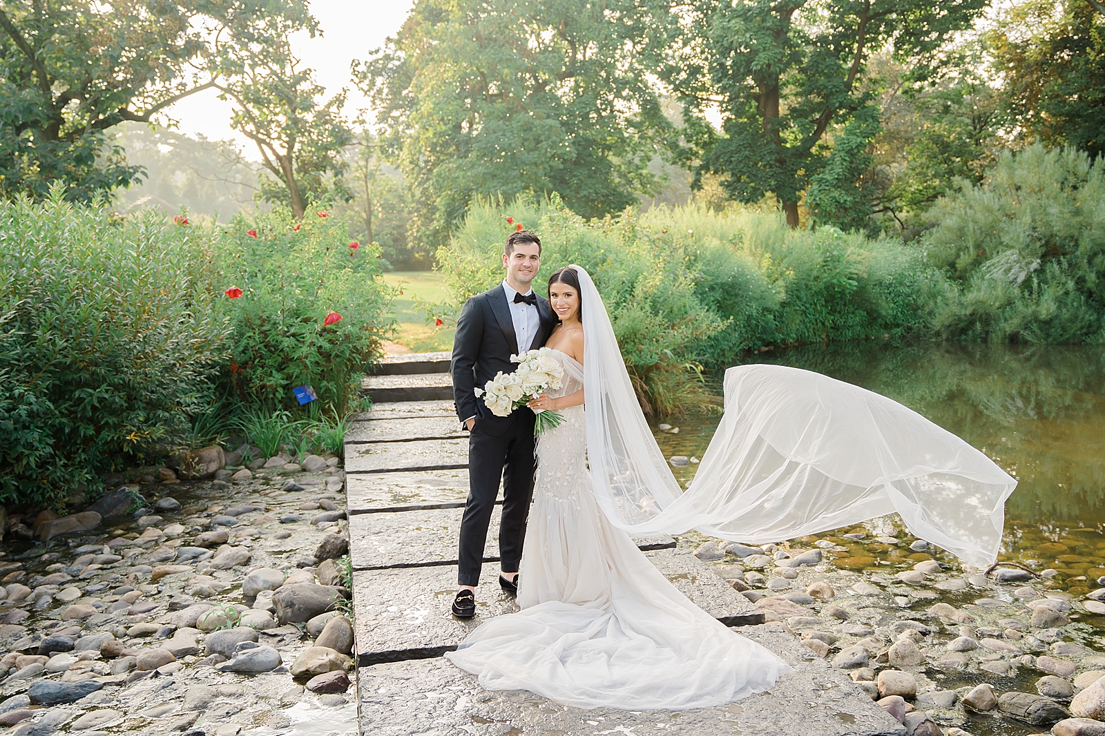 Full body shot of the bride and groom standing on a stone path that bisects a pond as they smile for the camera. 