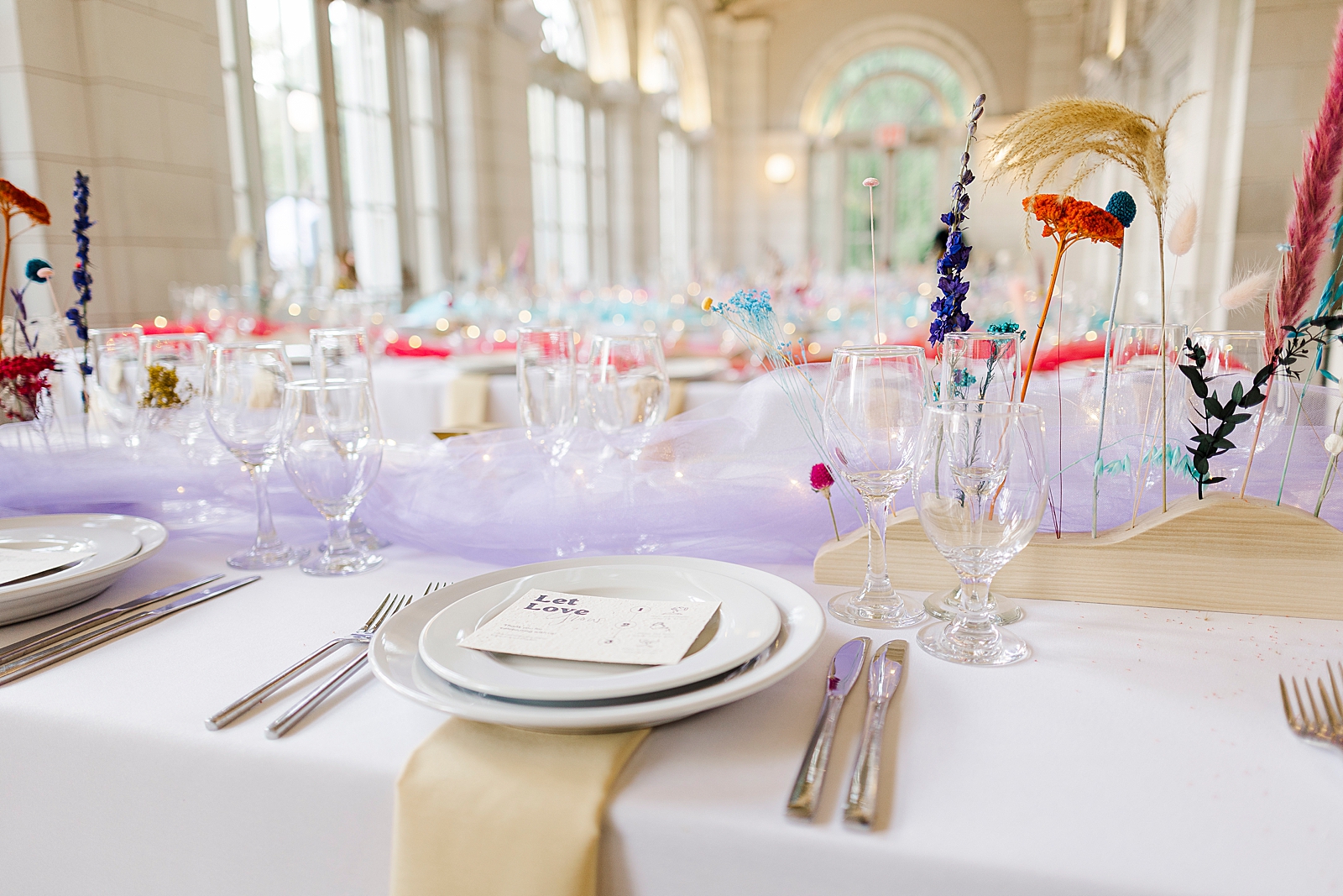 Shot of reception tables complete with glasses, plates, cutlery, floral arrangements, and colorful tulle centerpieces.