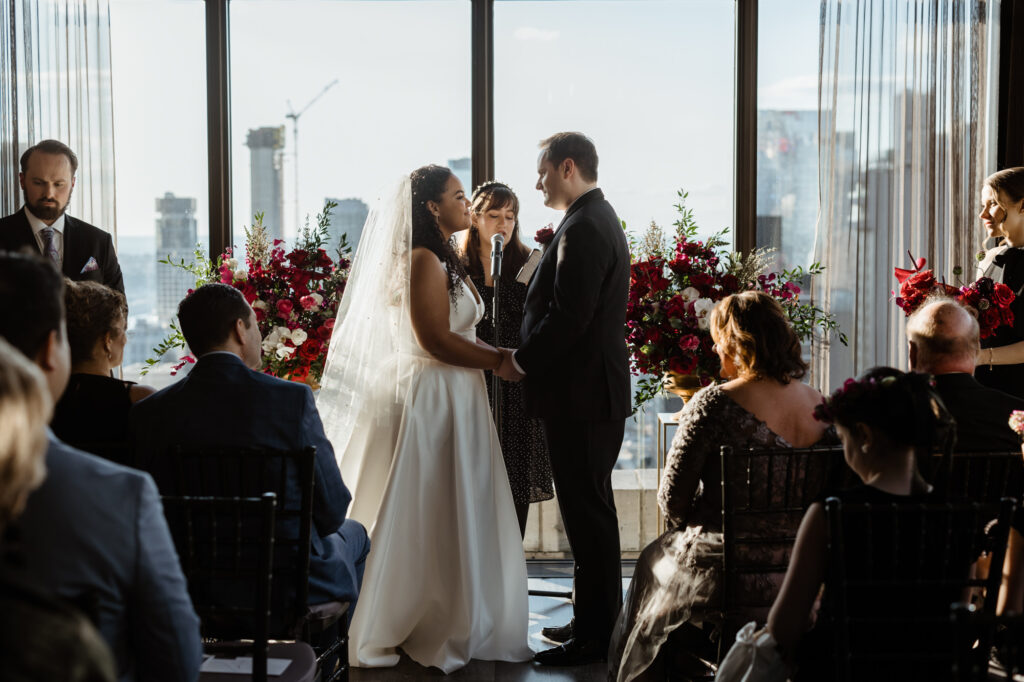Full body shot of a couple holding hands as they stand in front of a large window at the wedding altar. 
