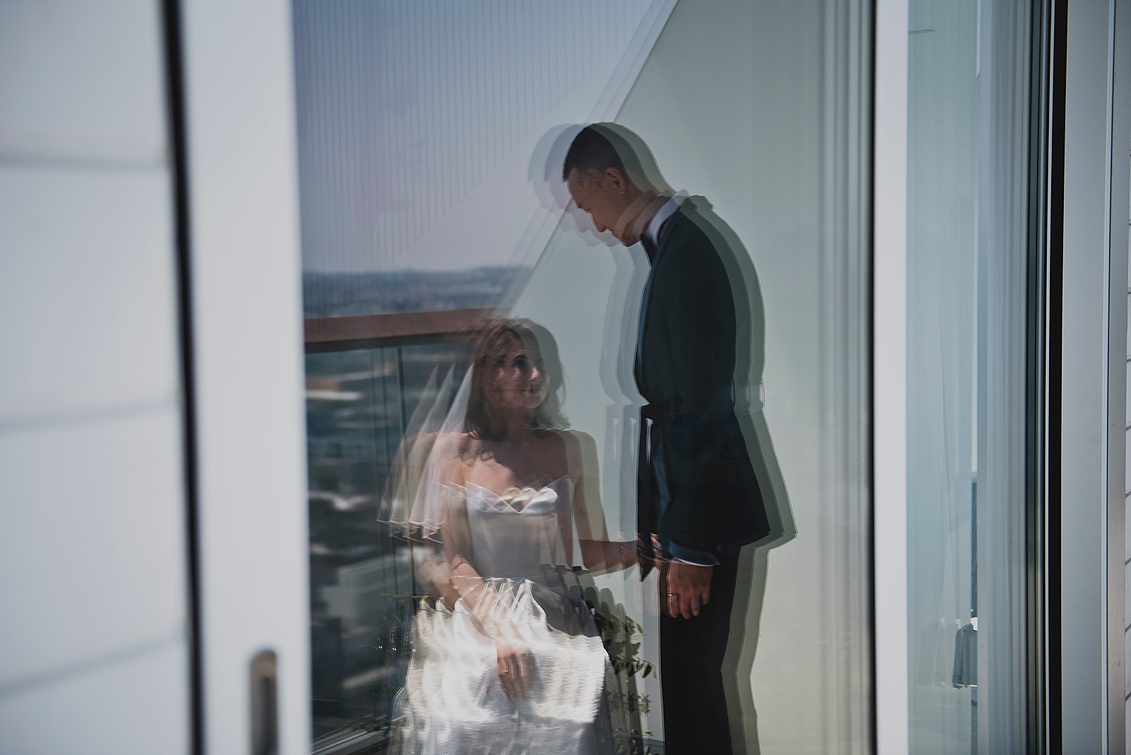 Shot of the bride and groom smiling at each other through the reflection of a glass door. 