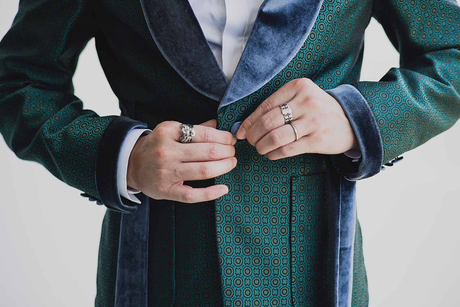 Close up shot of the groom buttoning his green and blue suit jacket. 