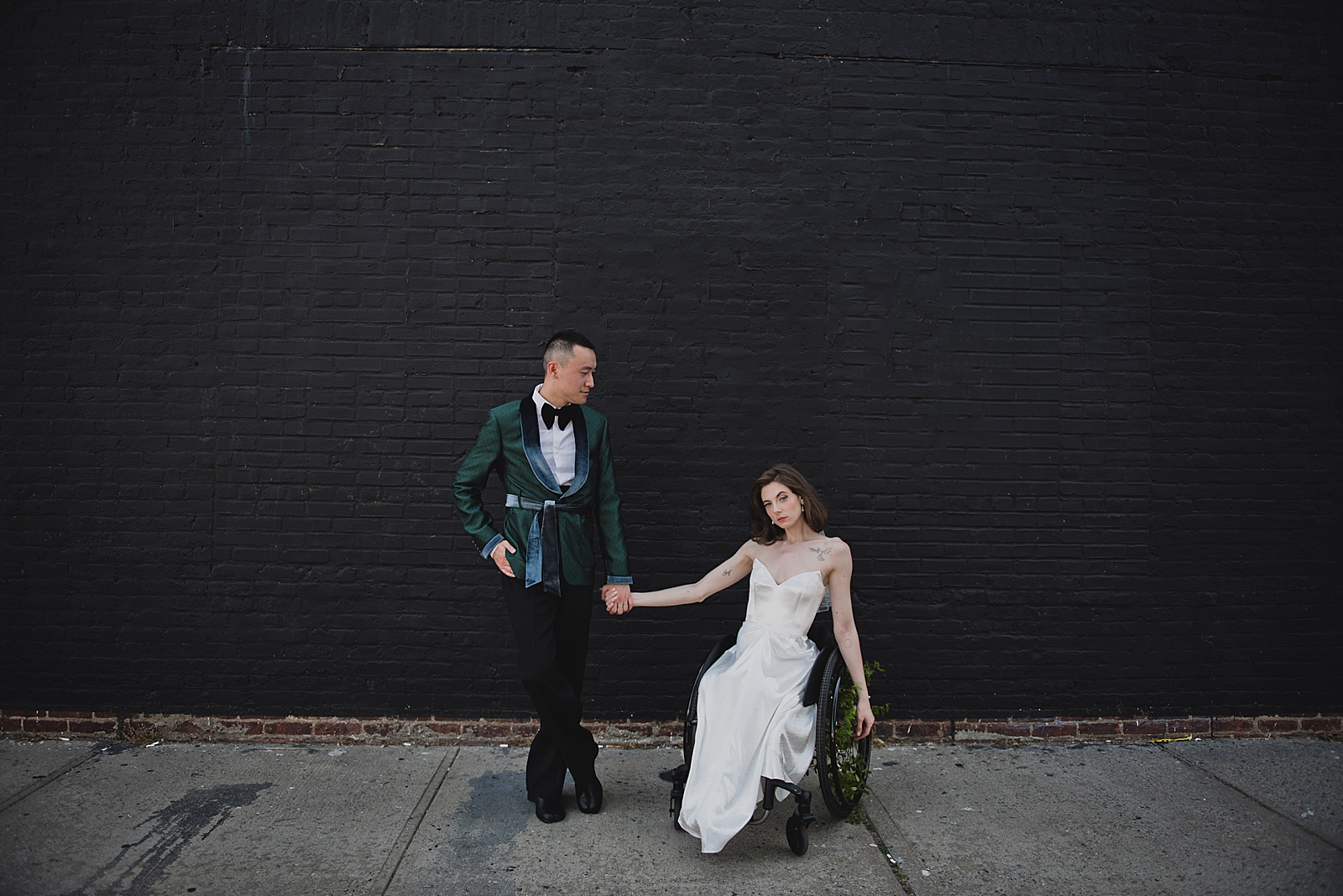 Full shot of the bride and groom posing in front of a black painted brick wall. 