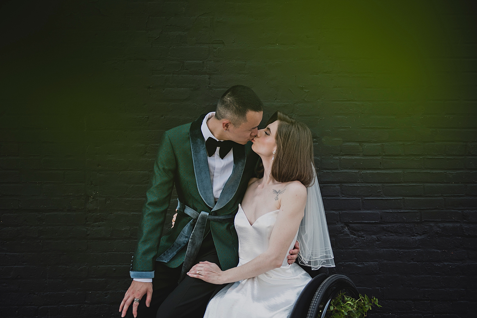 Upper body shot of the groom leaning on the bride's wheelchair as they share a kiss. 
