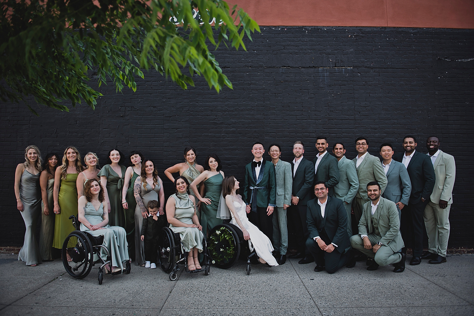 Full shot of the bride, groom, and bridal party as they pose in front of a black brick wall. 