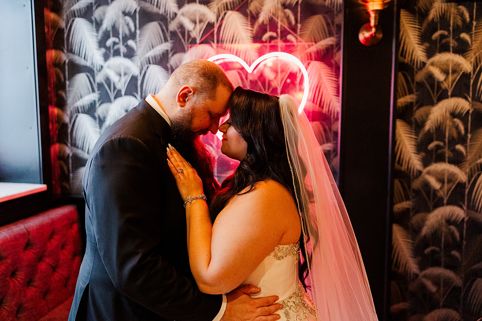 Upper body shot of the couple nose to nose as they embrace in front of a pink florescent light heart sign.
