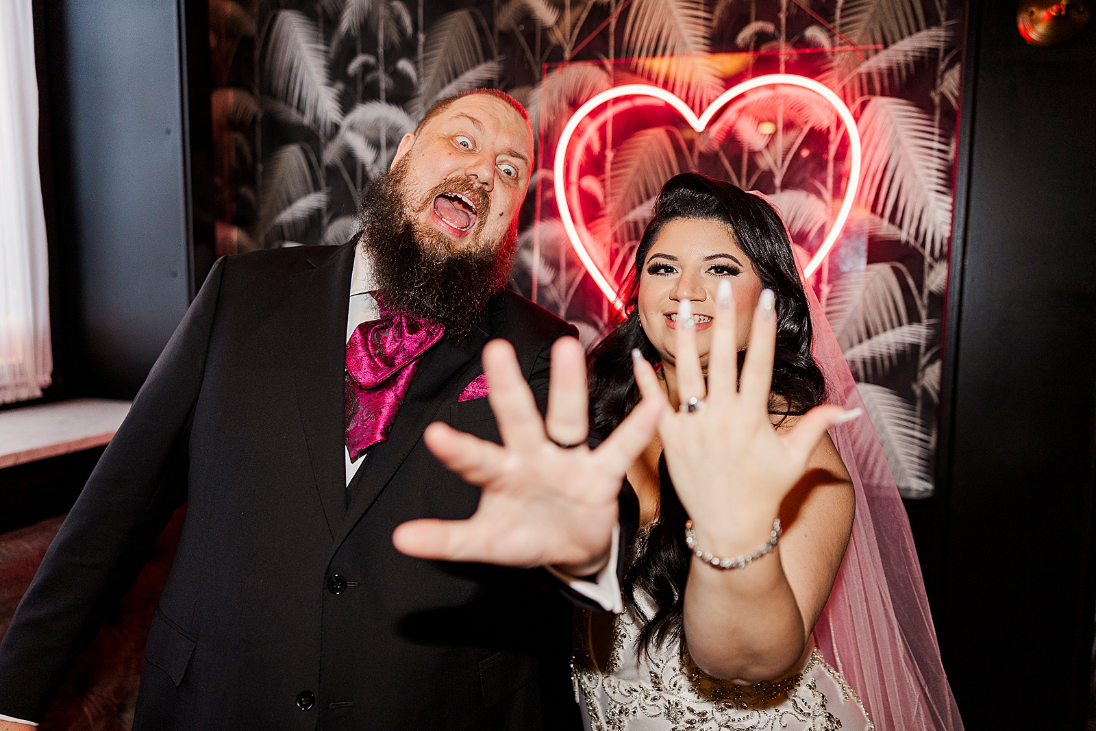Upper body shot of the couple showing off their rings in front of a pink florescent light heart sign.