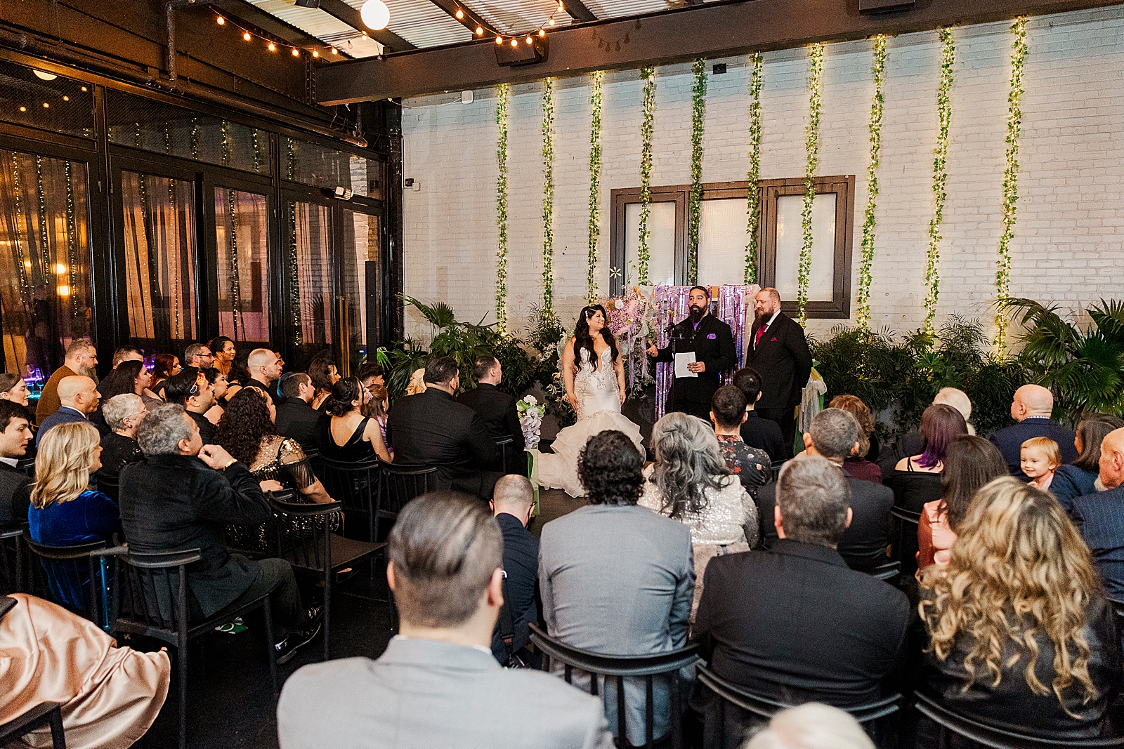 Full shot of the ceremony space, with guests in their seats and the bride and groom with the officiant standing at the altar. 
