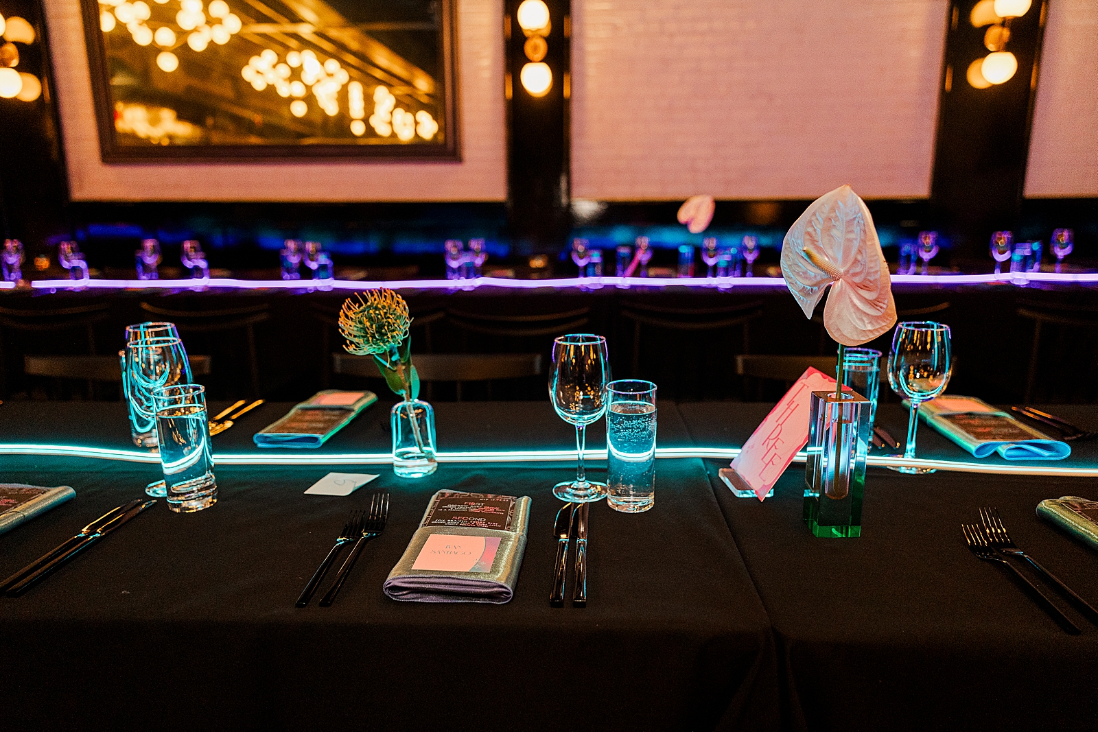 Close up shot of a table in the reception space, complete with menus, place cards, napkins, flowers, glasses, and a long neon light. 