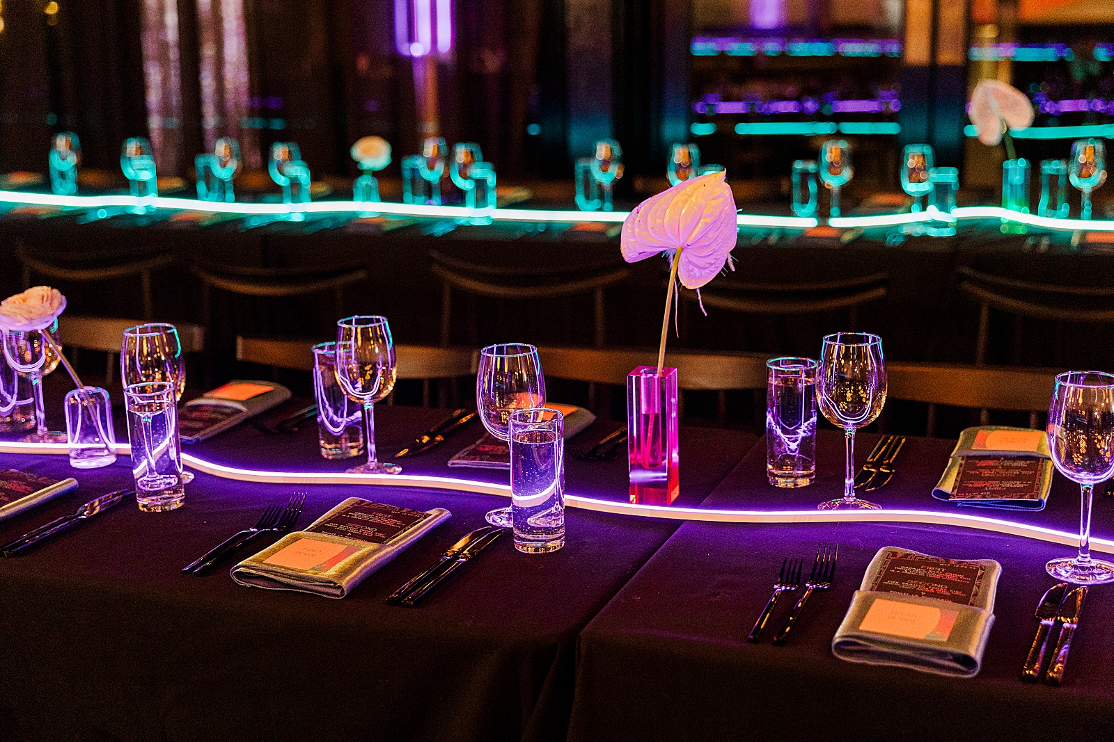 Close up shot of a table in the reception space, complete with menus, place cards, napkins, flowers, glasses, and a long neon light. 