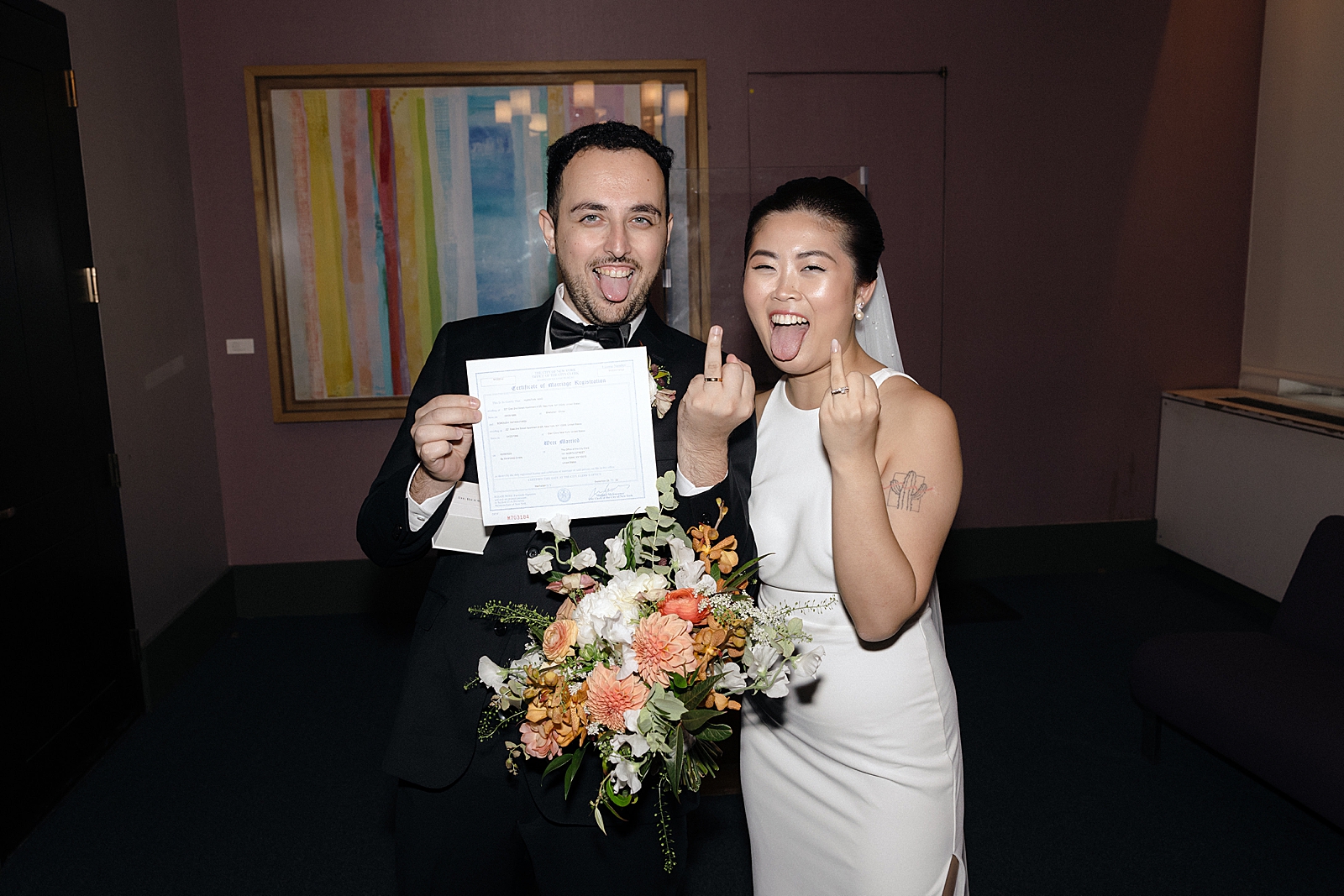 Upper body shot of the bride and groom posing with their ring fingers up as the groom holds their marriage certificate. 