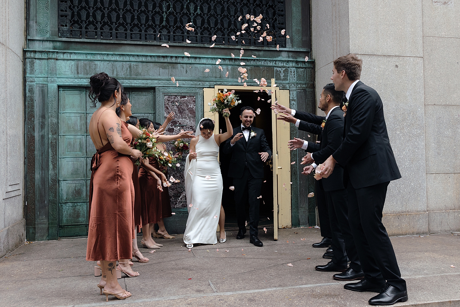 Full shot of the bride and groom exiting the courthouse as their bridal party showers them in flower petals. 
